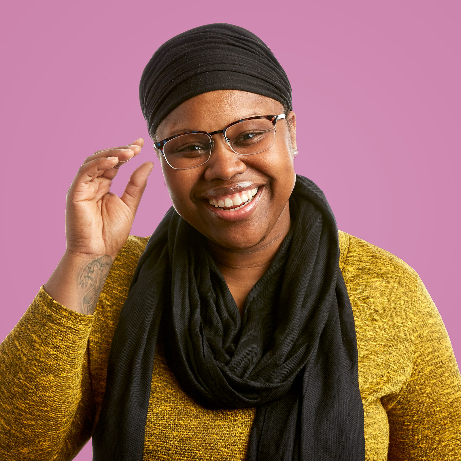 Person wearing black headwrap, glasses, a yellow sweater, and a black scarf, smiling against pink background.