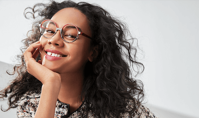 Woman wearing round, red and tortoiseshell eyeglasses, smiling with her hand on her cheek.