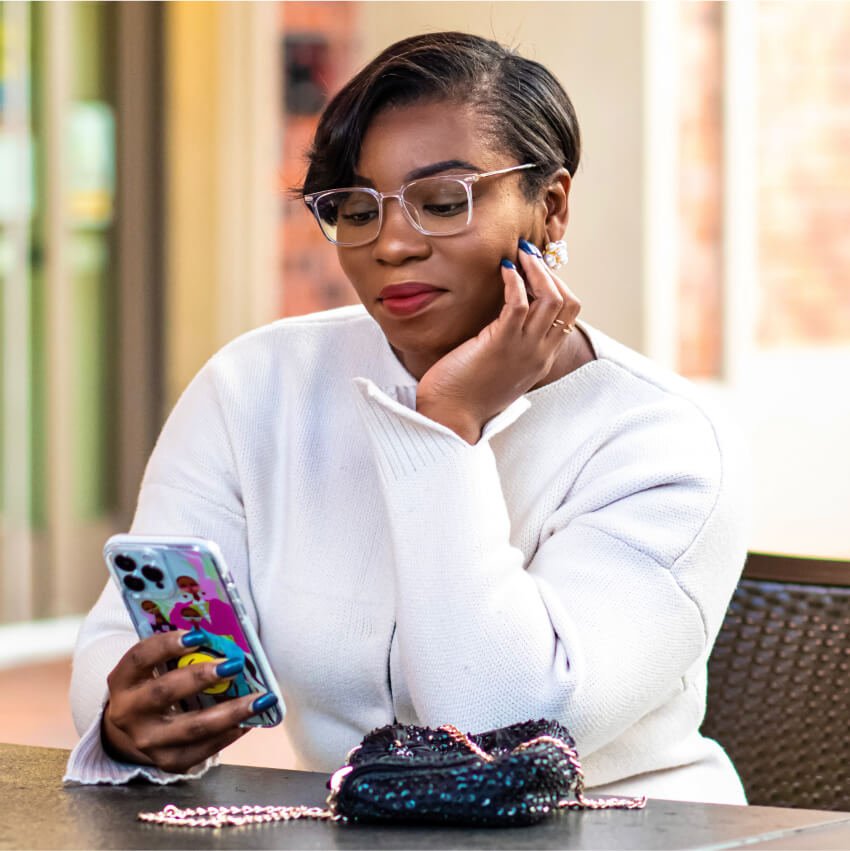 Person wearing glasses holding a smartphone with a colorful case, small black purse with chain on table.