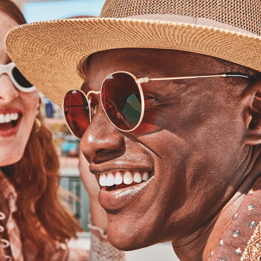 Man wearing a straw hat and round sunglasses with red lenses, smiling.