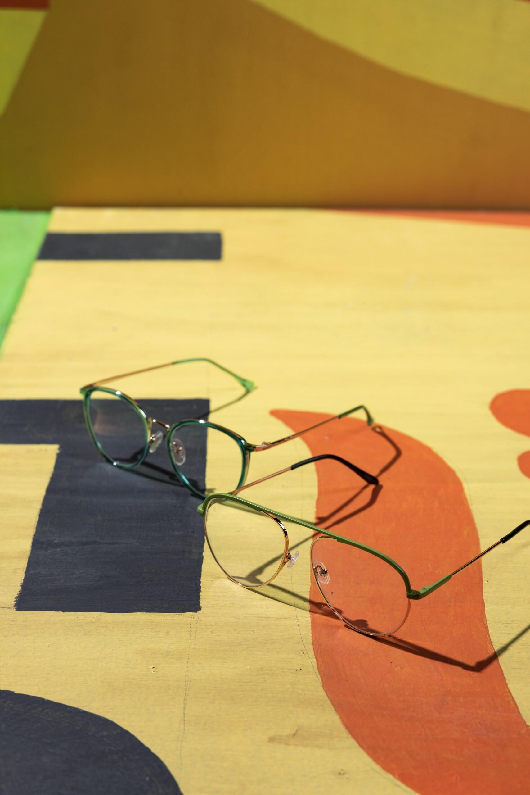 Two pairs of green-framed eyeglasses on a multicolored surface with abstract patterns.