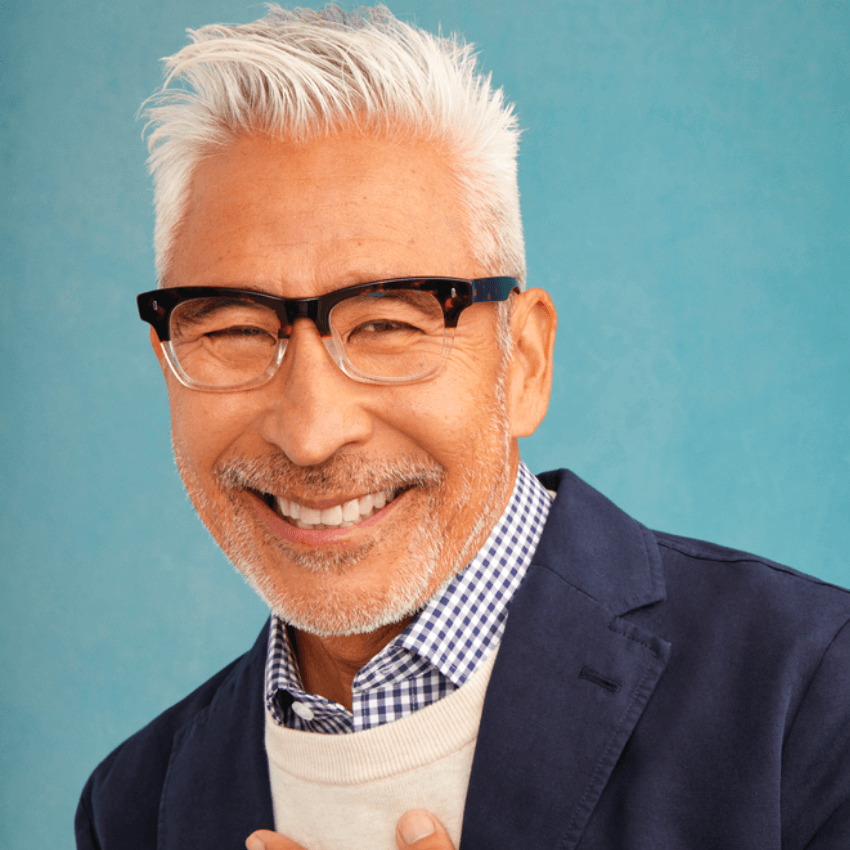 Man wearing stylish black-framed eyeglasses, light blue and white checkered shirt, and navy blazer, smiling.