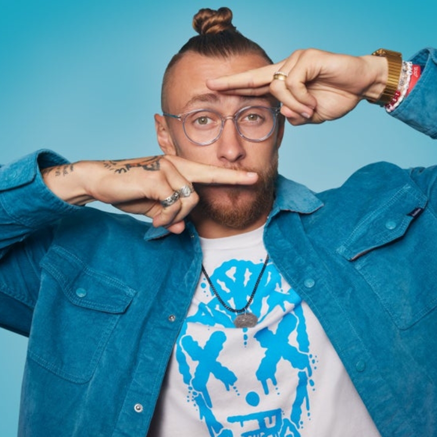 Man wearing glasses, a blue jacket, and a white t-shirt with a blue graphic design making hand gestures.