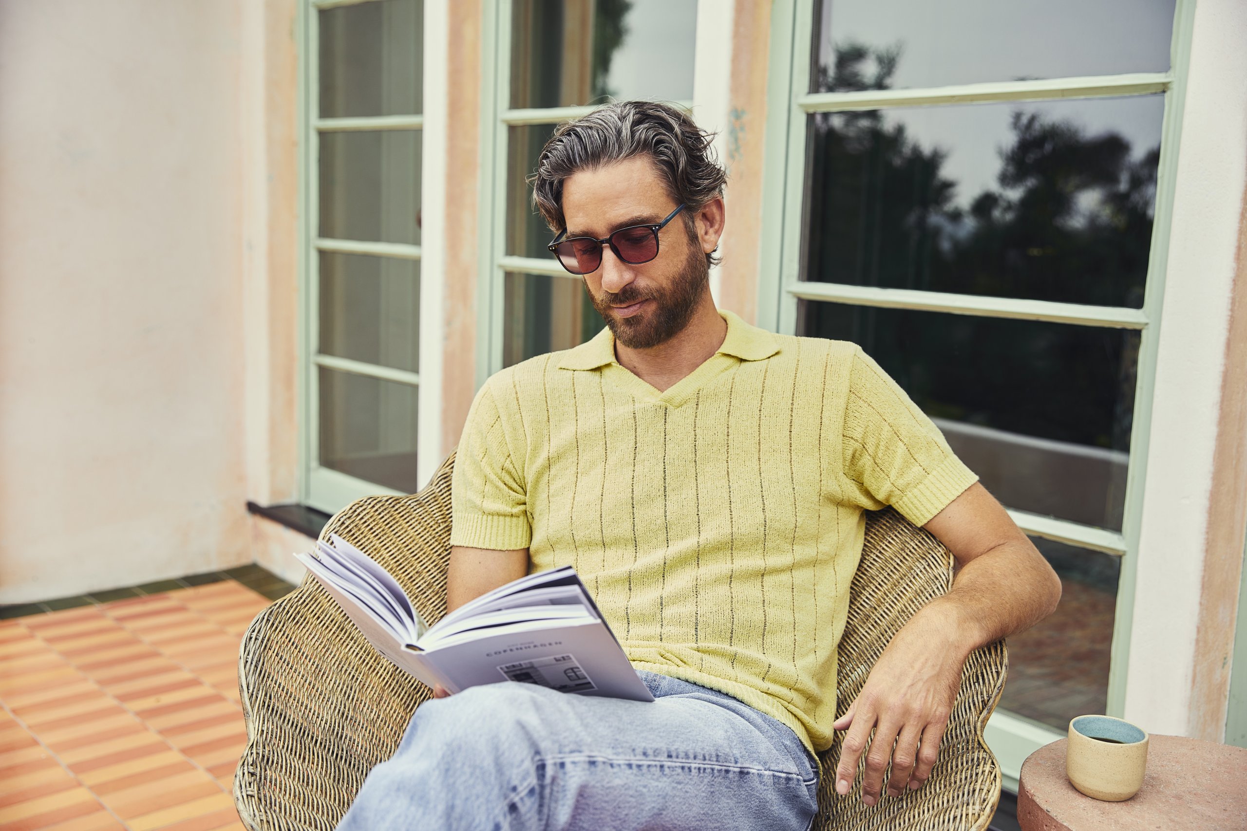 Man in yellow short-sleeve knit shirt and sunglasses, sitting in a wicker chair, reading a book.