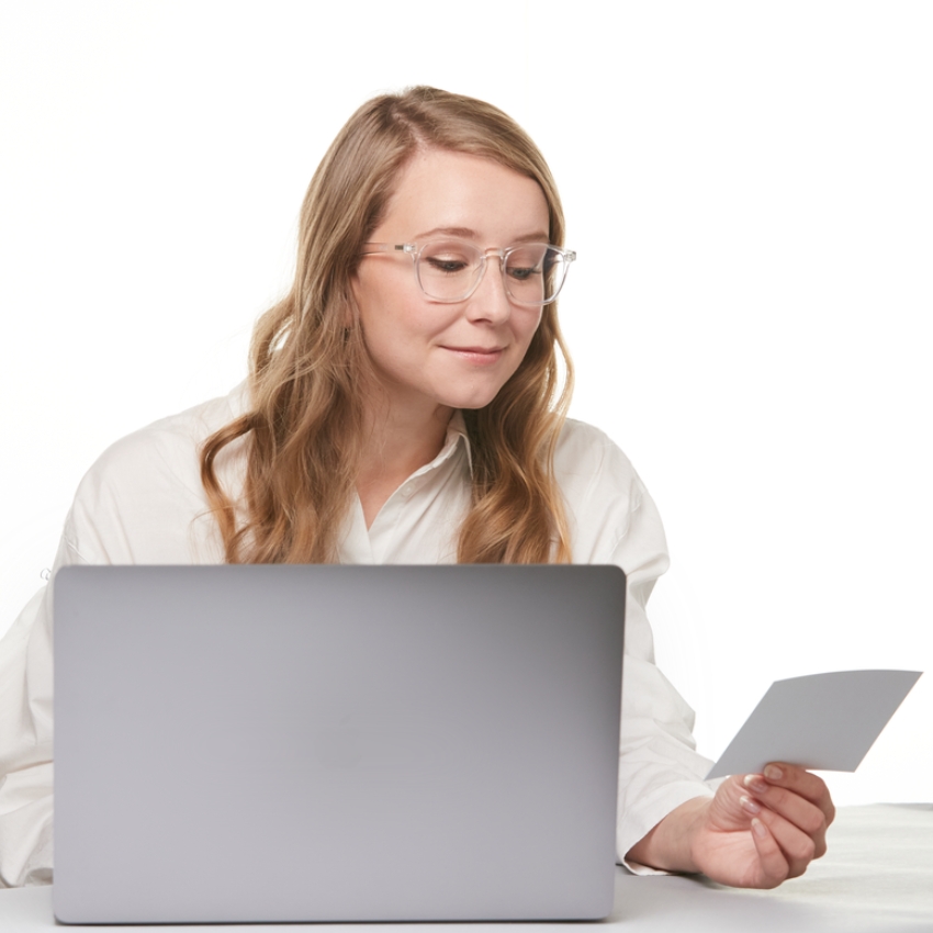 Person with glasses using a laptop while holding a piece of paper.