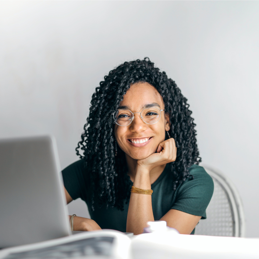 Smiling person with curly hair and glasses working on a laptop, resting chin on hand.