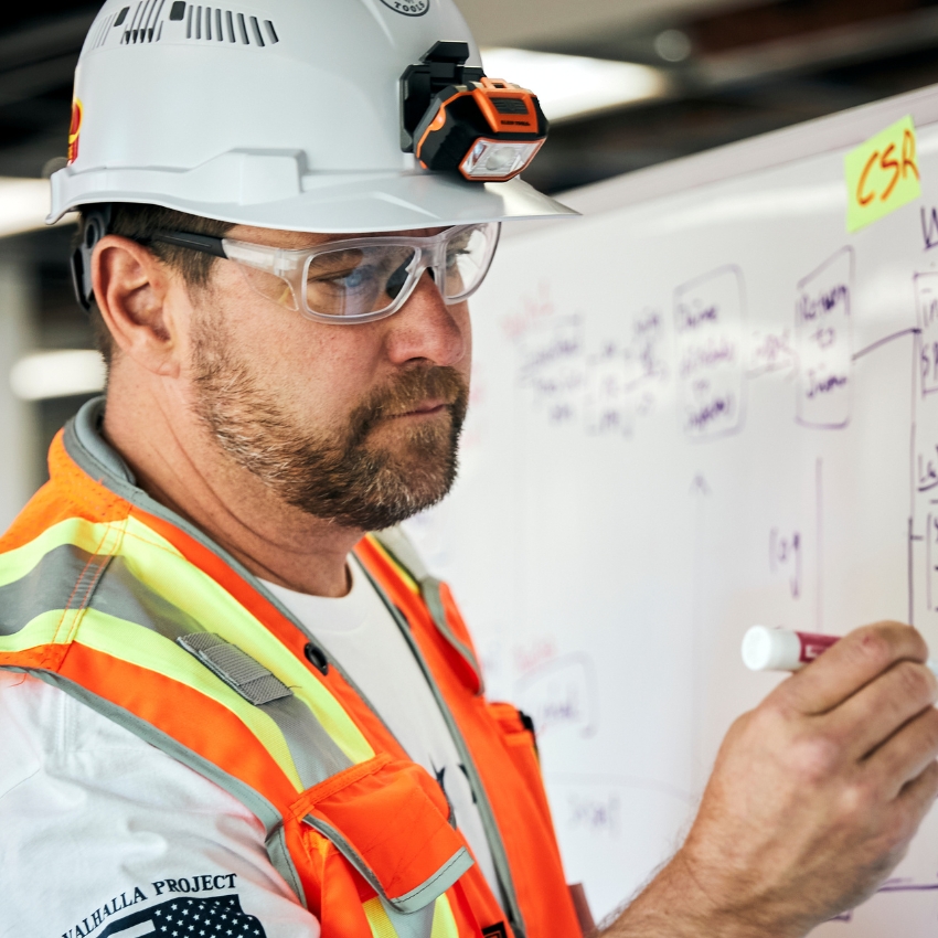 Worker wearing a white hard hat with headlamp, safety glasses, and a high-visibility safety vest writing on a whiteboard.