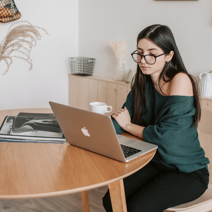 Woman working on a MacBook at a round wooden table with a magazine and a white mug.