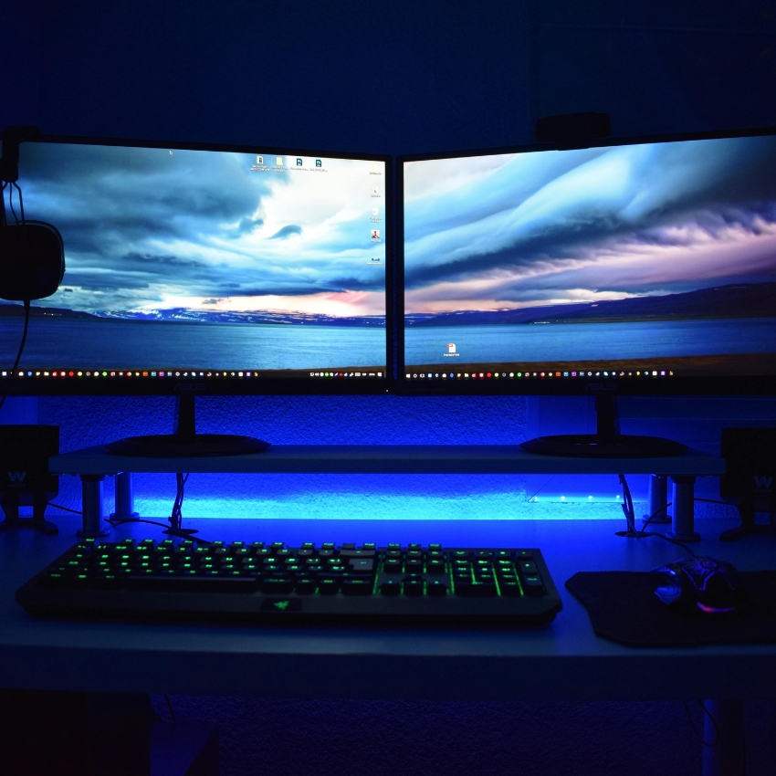 Dual computer monitors with a scenic ocean and sky wallpaper, backlit keyboard, and gaming mouse on mousepad.