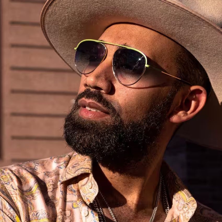 Man wearing a beige hat, aviator sunglasses with green frames, a floral patterned shirt, and necklace.