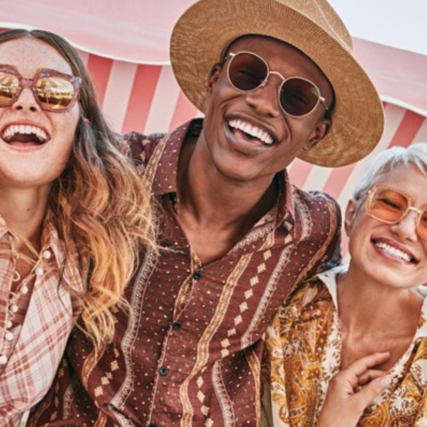 Three people wearing stylish sunglasses and patterned clothing, smiling and laughing together outdoors.