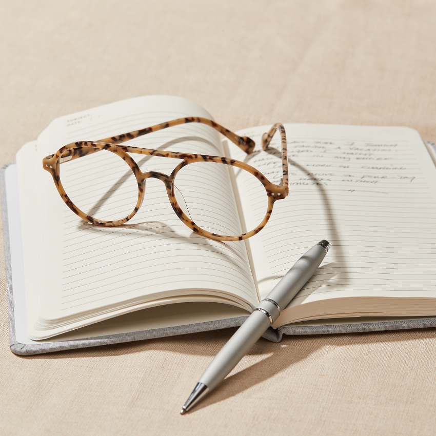 Tortoise-patterned eyeglasses and a silver pen resting on an open lined notebook.